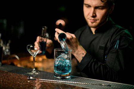 酒保把一杯蓝色的酒精饮料从盘车里倒到盛着玻璃瓶的量杯上