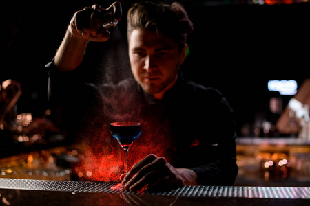 专业男调酒师在红灯下用苦涩的调酒师喷洒蓝色酒精鸡尾酒