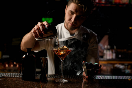 职业男调酒师用蒸汽将棕色酒精饮料从钢制摇壶倒到玻璃杯上