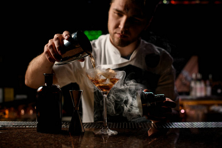 专业调酒师用蒸汽将棕色酒精饮料从钢制摇壶倒到玻璃杯上
