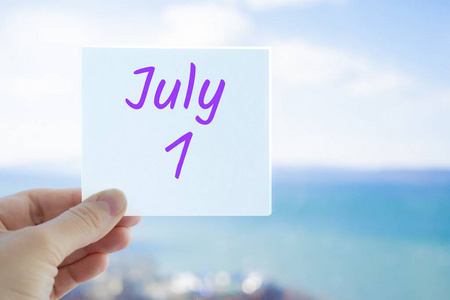 7月1日。手持贴纸，在模糊的海天背景上显示7月1日的文字。复制文本空间。月历概念