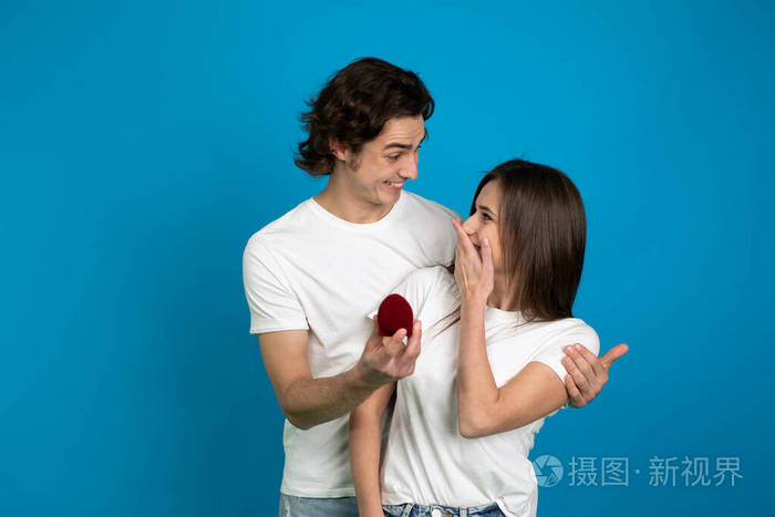 一个年轻人拥抱女友拿着蓝色背景上孤立的求婚戒指