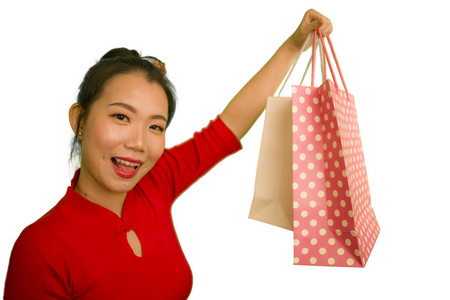 年轻美丽而兴奋的亚裔中国女子，穿着红色连衣裙，微笑着，兴奋地拿着购物袋，在时尚概念的白色背景下欢快地孤立着