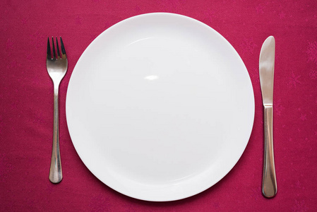 Empty white plate, fork and knife on maroon tablecloth. Elegant 