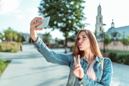 穿着牛仔裤的女孩,夏天在城市公园,手里拿着智能手机,在网上拍照,自拍