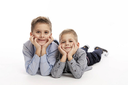 两个穿蓝色衬衫的男孩兄弟躺在地上，双手托着头，微笑着。白色背景。