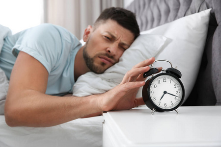 Sleepy man turning off alarm clock at home in morning, focus on 