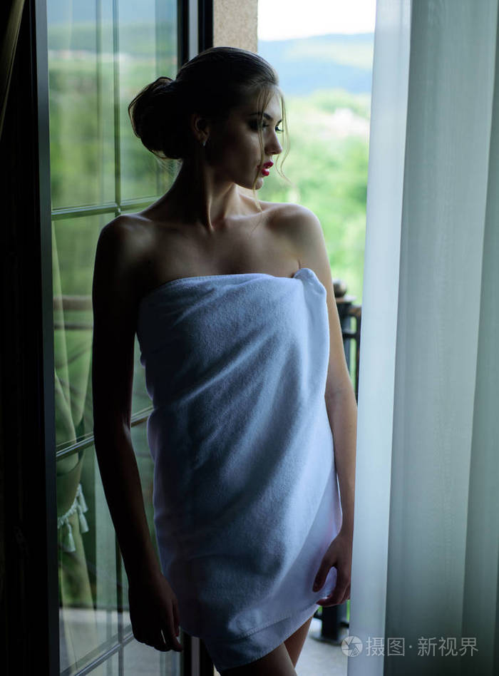 洗完澡后裹着毛巾的性感年轻女子站在窗前向外张望。一个漂亮的女人，在橱窗的背景下有着奢华的妆容和发型。