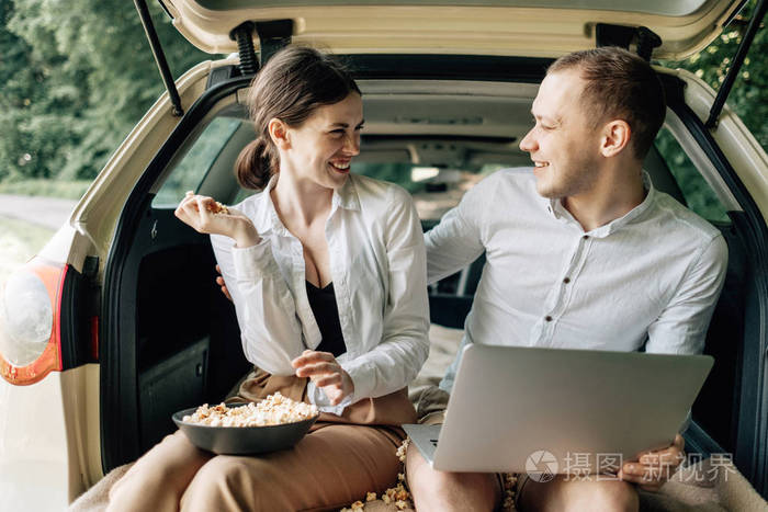 年轻幸福的情侣穿着同样的白色T恤坐在汽车后备箱里，在路边放着笔记本电脑和爆米花，周末出城，度假和公路旅行的概念