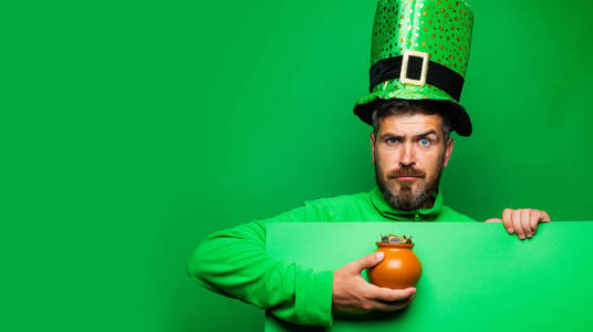 戴着绿帽和连帽衫的小妖精拿着一罐金子。一个留着胡子的男人，穿着小妖精的服装，背景是绿色的。大胡子男人庆祝圣帕特里克节。