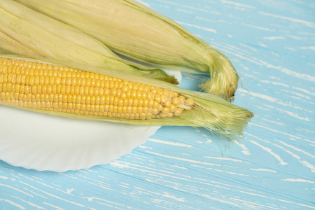 Corn cob with green leaves lies on white plate blue color backgr