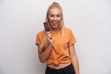 有趣的照片是一个快乐的年轻的金发女人穿着橙色的t恤，在白色的背景下摆着冰淇淋摆出愉快的舌头对着镜头