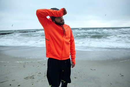 这是一个活跃的年轻黑发胡子运动员，在海边晨练后，用他温暖的橙色运动外套袖子擦汗