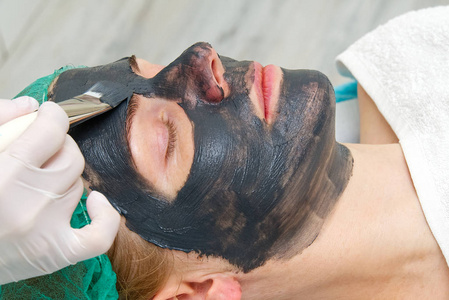 美容师将天然保湿面膜涂在年轻美女的脸上。黑竹炭面膜，用刷子涂在脸上。