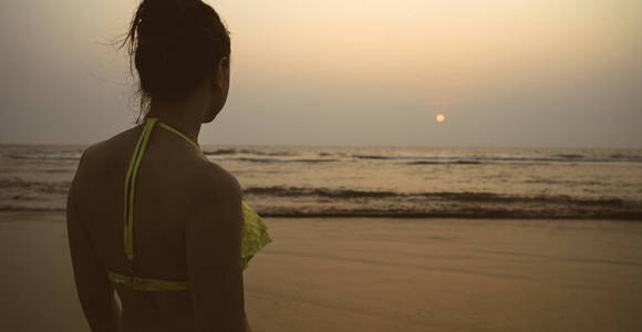 日落时海边的无名女子。傍晚在度假胜地波涛汹涌的大海附近的海滩上站着一个不知名的穿着泳装的女性的后视图