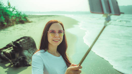 快乐的女人在海边自拍。从上面看，一个快乐的女人在沙滩上的波涛汹涌的海面上用单脚架和智能手机自拍