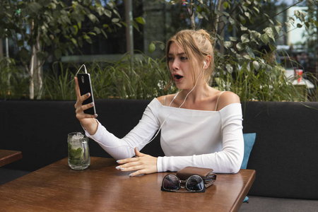 喝鸡尾酒的金发女博主惊呆了，她正在咖啡厅通过连接免费wifi的智能手机阅读收到的短信。积极的年轻学生在现代手机上观看在线广播。