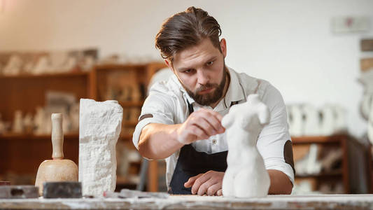 这位留着胡须的雕塑家在创意工作室测量石头女人的躯干,从石灰石中