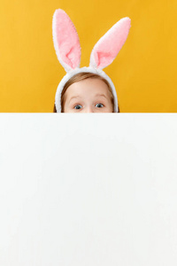 复活节快乐。一个长着兔子耳朵的有趣的小女孩因为一条空白的横幅而躲藏起来。这孩子吃鸡蛋