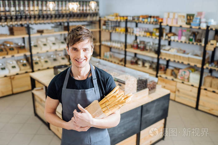 穿着灰色围裙的专业白种面包店老板抱着grissini微笑着邀请客人来自己
