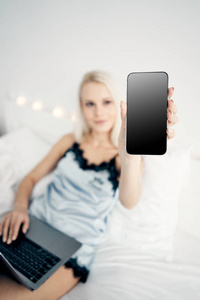 美丽的年轻女孩坐在床上，展示她的智能手机屏幕。