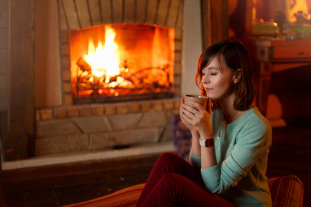 女人在壁炉边喝茶取暖.年轻的白人女性在家里端着一杯咖啡.