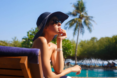 年轻女性戴着草帽，在时髦的海滩上享受热带阳光，坐在躺椅上。夏季护肤理念。美丽的女孩在海边的岛上度假