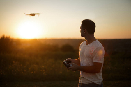 日落时，一个手拿遥控器在空中驾驶无人机的人的侧影。飞行员在空中用四旋翼机拍摄空中照片和视频