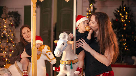 圣诞概念两个婴儿坐在室内旋转木马上和他们玩一个婴儿嚼着圣诞球他的妈妈吻他的脸颊