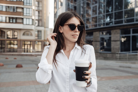 一个穿着牛仔裤和白衬衫的时髦女孩喝着咖啡，用她的智能手机，商务小姐，女人的力量概念