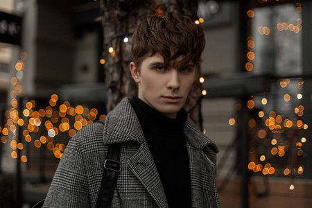 一个年轻人穿着优雅的外套，背着一个老式的皮革背包，在一家装饰着明亮新年灯光的咖啡馆附近。穿着时髦衣服的帅哥时尚模特正在城市里散步