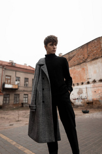 年轻的欧洲男模，一个时尚的发型，灰色格子外套，牛仔裤，黑色复古毛衣，站在外面的街道上的建筑物之间。不错的时髦的欧洲人。