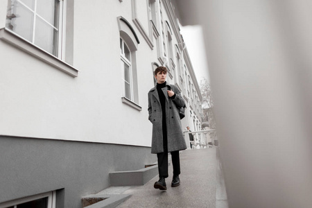 一个欧洲年轻人穿着时髦的格子灰色外套，穿着黑色时髦的靴子，背着一个时髦的皮包，走在靠近墙的户外。时髦迷人，穿着优雅衣服的现代人。