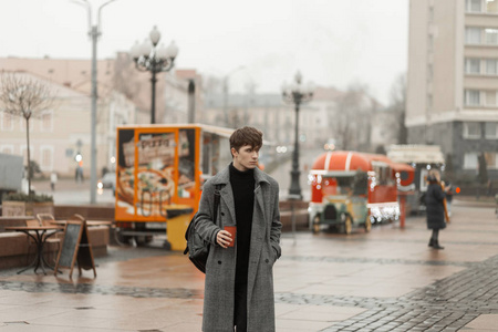 一个年轻人穿着时髦的格子冬季外套，穿着一件复古的黑色毛衣，在街上喝着热腾腾的咖啡。一个欧洲人带着美味的饮料在城市里四处旅行