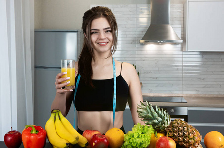 身材修长，圆领，穿着黑色运动上衣，站在装满水果的厨房里，拿着一杯橙汁营养学和营养学的玻璃杯