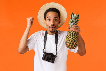 万岁假期和热带水果。快乐兴奋的非洲裔美国人终于出国旅行了，旅途中玩得很开心，拳头打气，手里拿着菠萝，站在橙色背景上的相机前