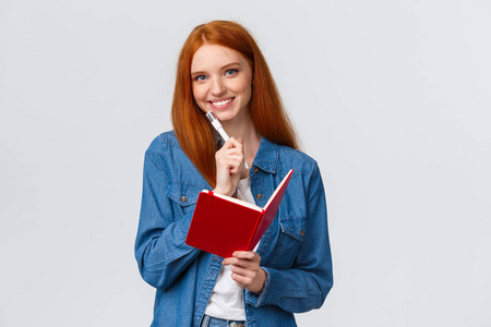创意可爱帅气红发女学生，十几岁穿休闲装，拿着钢笔和微笑的相机，在笔记本上记笔记，写日记，准备时间表站在白色背景下