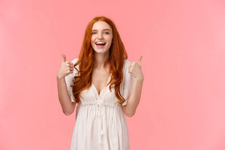 腰间肖像开朗，乐观的红发女子身穿白色连衣裙，竖起大拇指，微笑着表示赞同，喜欢产品，推荐它，表达积极的意见，站在粉红色的背景上