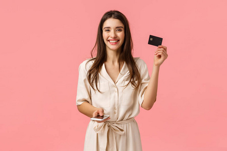 广告技术和数字生活方式概念。无忧无虑迷人的年轻女子穿着华丽的衣服，出示信用卡，手持智能手机，微笑着在网上购物，粉色背景