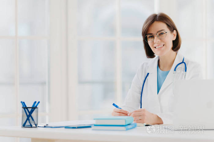 医生穿着白色长袍，带着听诊器，在医院工作，做笔记，在自己的柜子前摆姿势，坐在桌子旁，办公室里的笔记本电脑开心地笑着。保健与医疗理