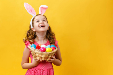 美丽可爱的小女孩戴着复活节兔子耳朵，手里拿着一个黄色背景上有鸡蛋的篮子。那孩子抬起头，抬起头来。复制空间