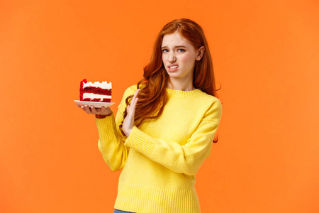 厌恶和不高兴，挑剔的红发女孩注意身材，不吃糖果，拿着块好吃的蛋糕拒绝吃，显示停止标志，厌恶地对着镜头做鬼脸，橙色背景