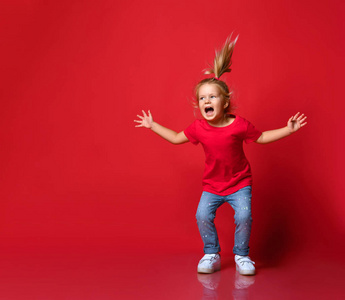 穿着时尚休闲服和白色运动鞋的小快乐兴奋的金发女孩在红墙背景上跳跃和感觉好玩