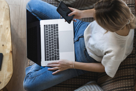 一位女士拿着笔记本电脑坐在沙发上用智能手机。在线学习和工作，自由职业者。一个个体户女孩坐在沙发上，桌上放着电话和电子书阅读器，手