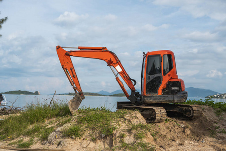 一台橙色的小型挖掘机在蓝天碧海的地面上进行施工。站在背景是蓝色海洋的地面上的小型履带式挖掘机。重工业。