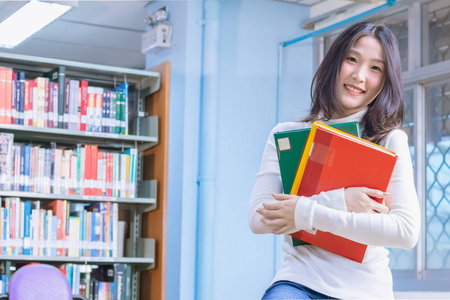 年轻的亚洲美丽微笑女学生肖像坐在和幸福的教科书在大学图书馆书架背景。教育知识和学习观念。