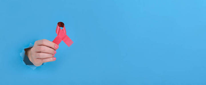 红丝带是抵抗艾滋病或女性乳腺癌疾病的象征。女孩的手拿着一条红丝带，背景是蓝色的碎纸纸。为你的广告内容复制空间