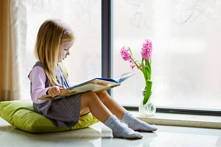 可爱的金发小女孩坐在阳台的地板上看书。教育爱好休闲家庭学校理念