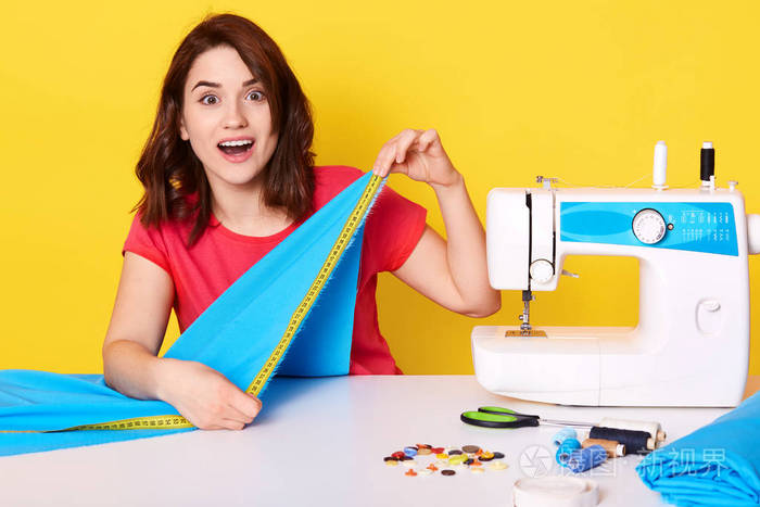 开心惊讶的女人用缝纫机做衣服,用卷尺,缝制新收集的衣服,坐在桌边