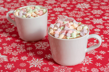 白色的热巧克力饮料杯和彩色棉花糖放在红布桌上，上面放着雪花